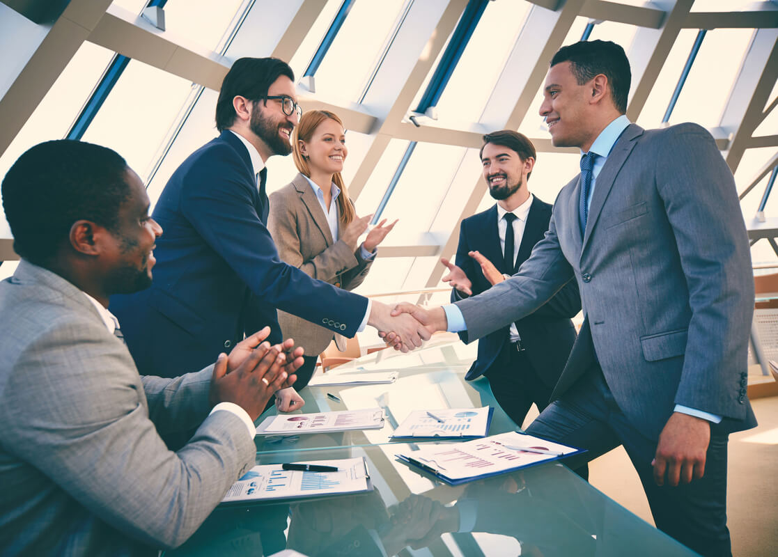 Five diverse business professionals in suits celebrating a successful deal with handshakes and applause in a modern conference room with large windows, business documents and charts visible on the glass table