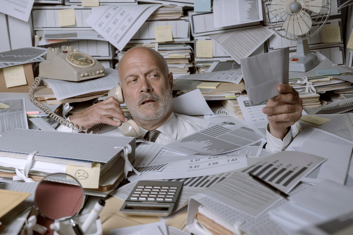 A man, overwhelmed, is surrounded by towering stacks of paperwork and office supplies. He holds a phone in one hand and examines a document with a weary expression.