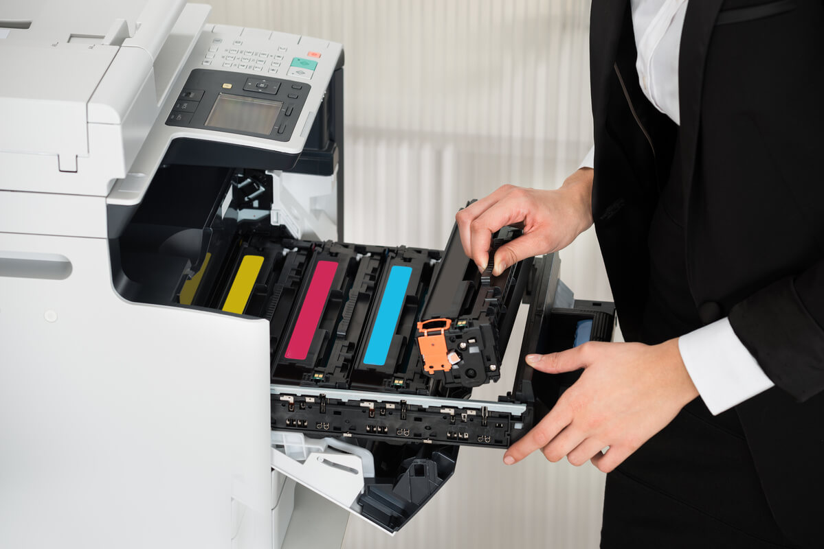 A person in business attire is replacing a black toner cartridge in an office printer, which has open slots for yellow, magenta, and cyan cartridges.