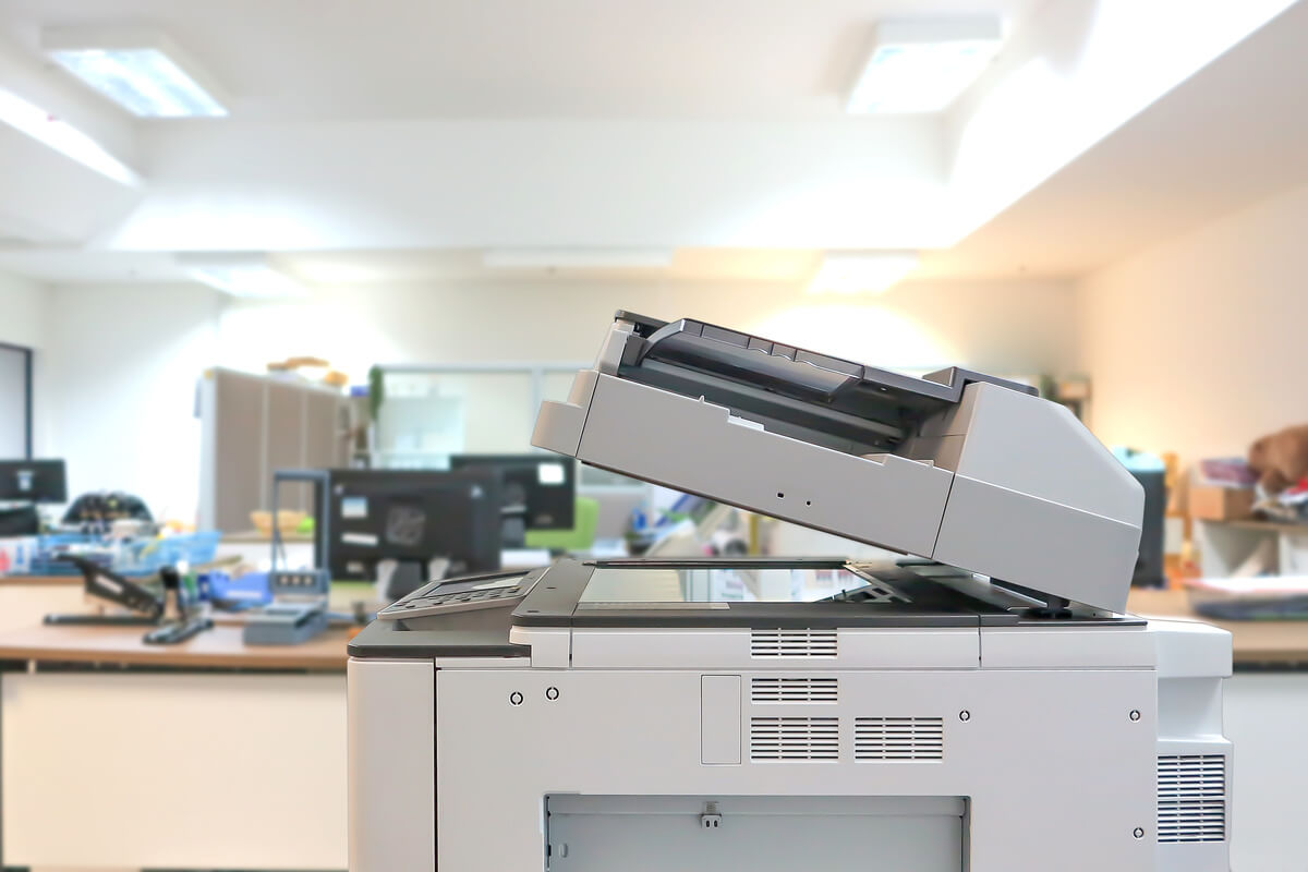 A photocopier with an open lid in a bright, modern office setting. The background is blurred, showing desks and office equipment, conveying a busy workspace.