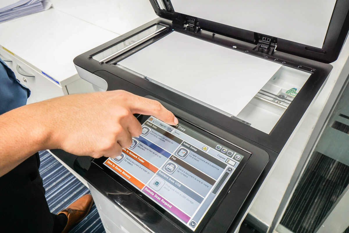 A person uses a touchscreen on an open office printer-scanner. The screen displays various options, indicating a task selection. The setting is modern and professional.