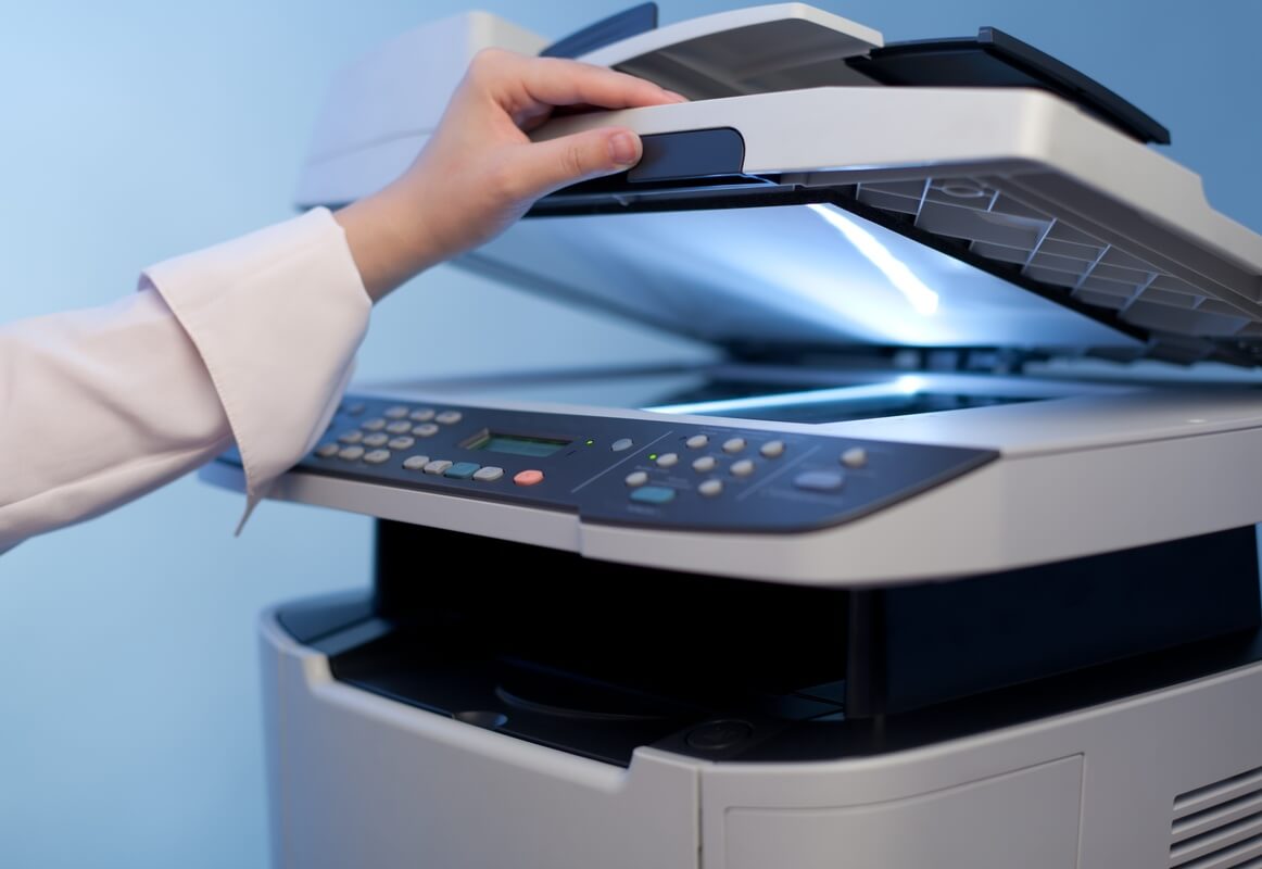 A person's hand lifts the lid of a photocopier, revealing a bright scanning light. The machine's control panel shows buttons and a small outdated display screen.