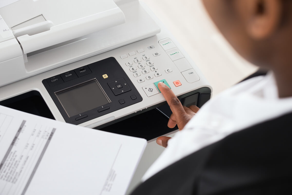 Close-up of a person operating a copier, pressing buttons on the control panel with one hand and holding documents with the other. 