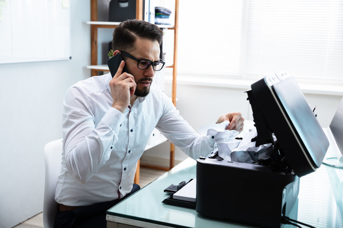 Man in an office, wearing glasses and a white shirt, on the phone while fixing a jammed printer. He looks frustrated, papers are crumpled around.