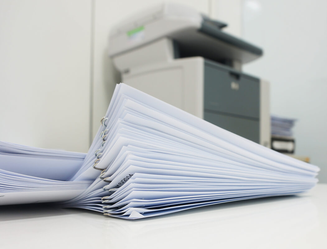 A large stack of white papers clipped together rests on a glossy desk. A gray office copier is blurred in the background.