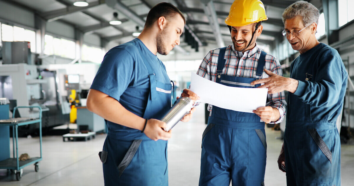 Three factory workers in blue overalls review blueprints.