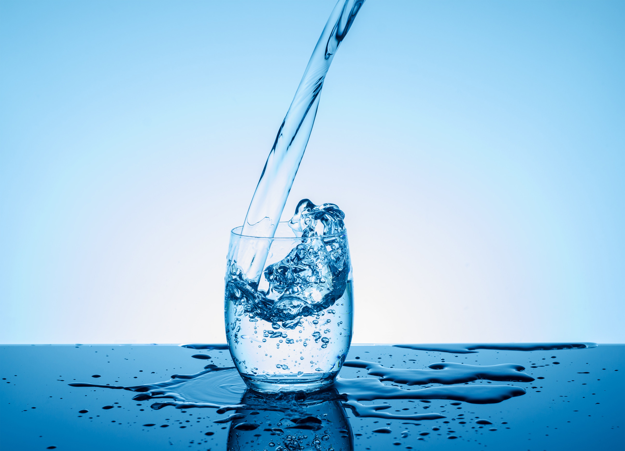 A glass is being filled with water against a blue background. Water splashes over the rim, creating ripples on the reflective surface below.