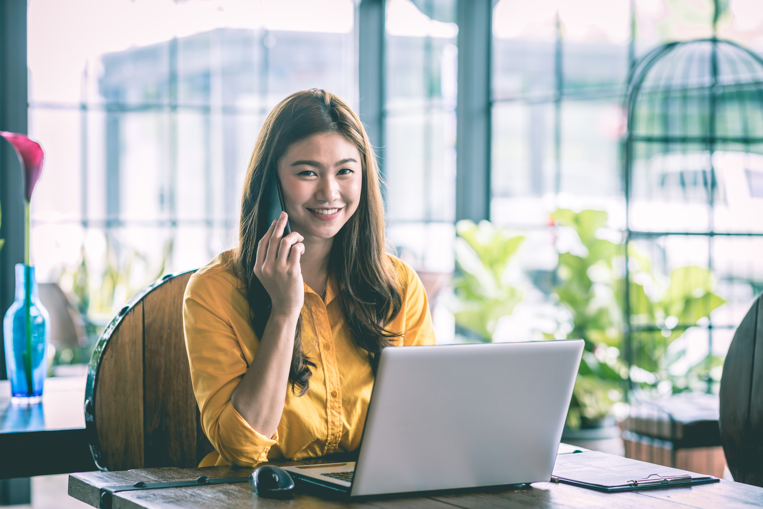 Smiling woman talking on phone while working at laptop in bright office