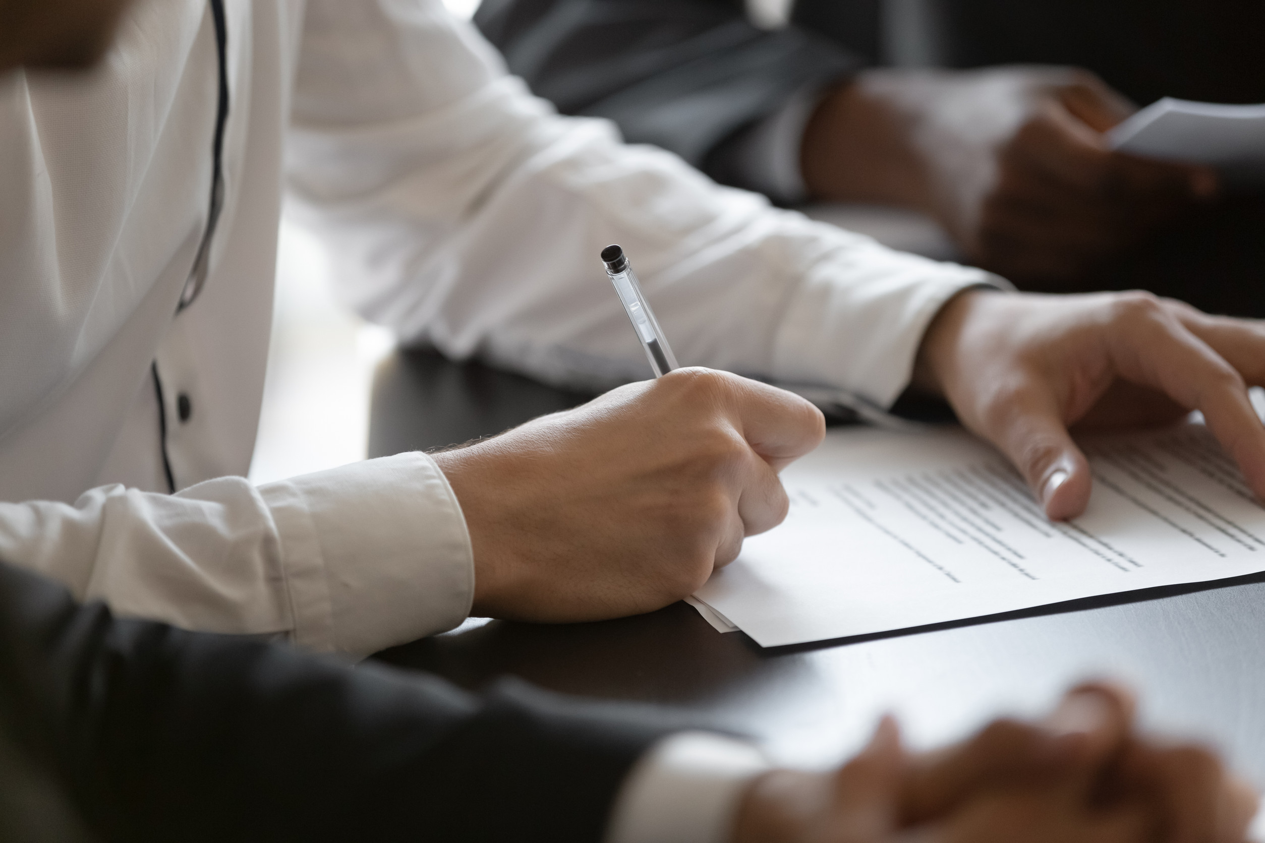 A person in a white shirt writes with a pen on a document at a dark table.