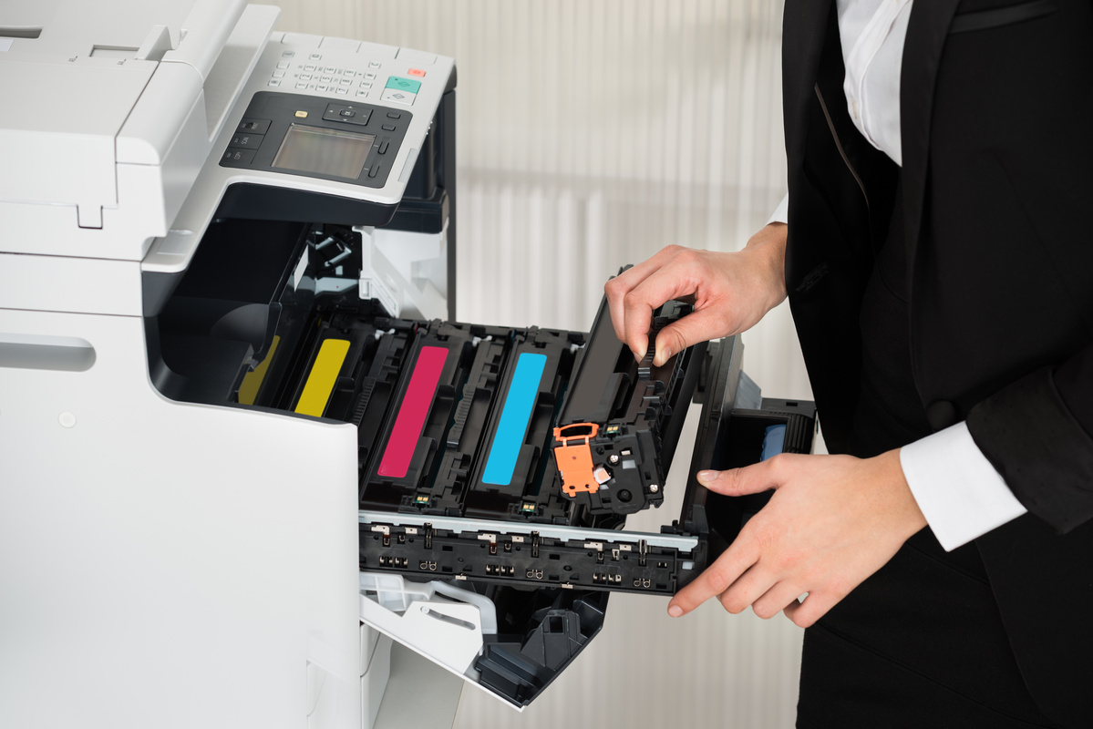 A person in business attire replaces a cartridge in an office copier. The open machine shows cyan, magenta, yellow, and black cartridges. 