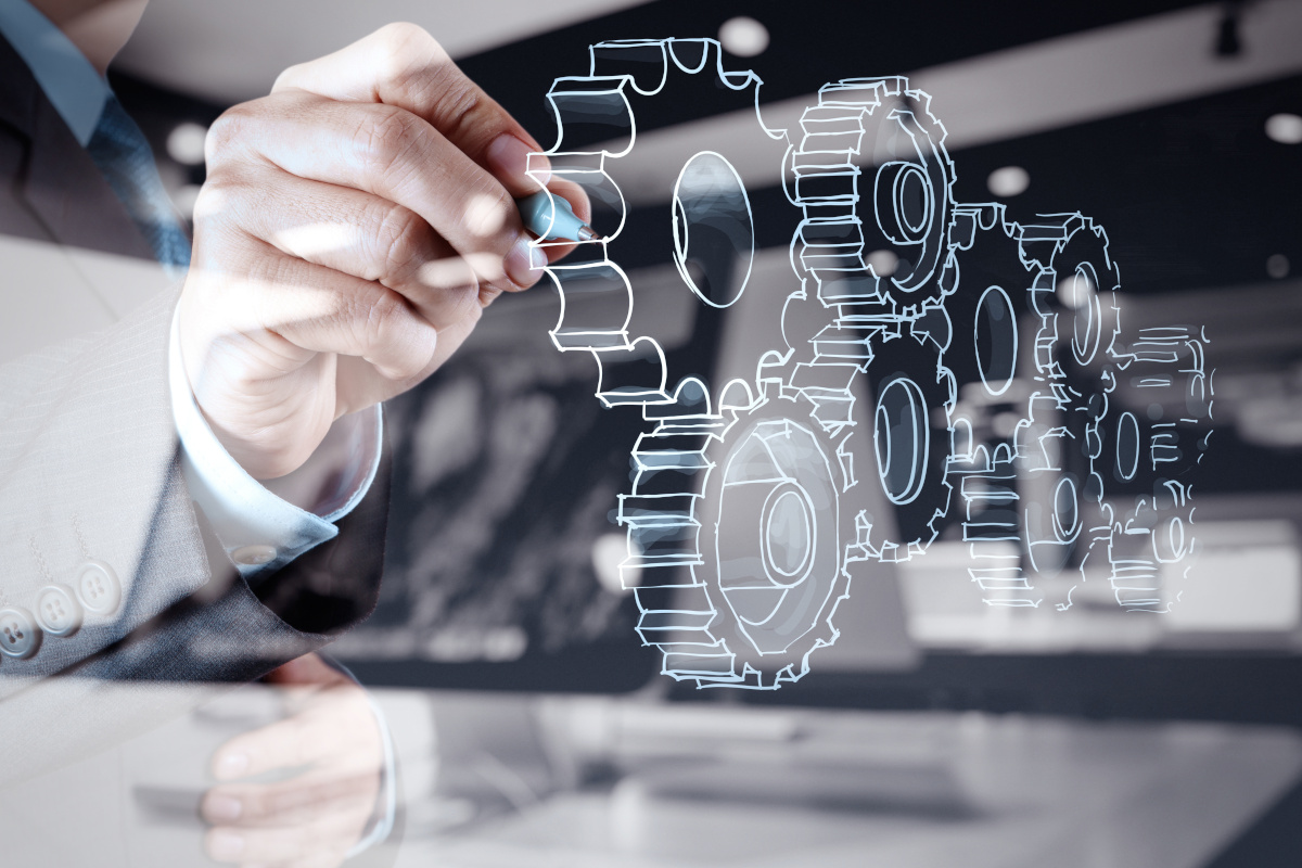 A person in a suit draws interconnected gears on clear glass with a marker, symbolizing teamwork and engineering.
