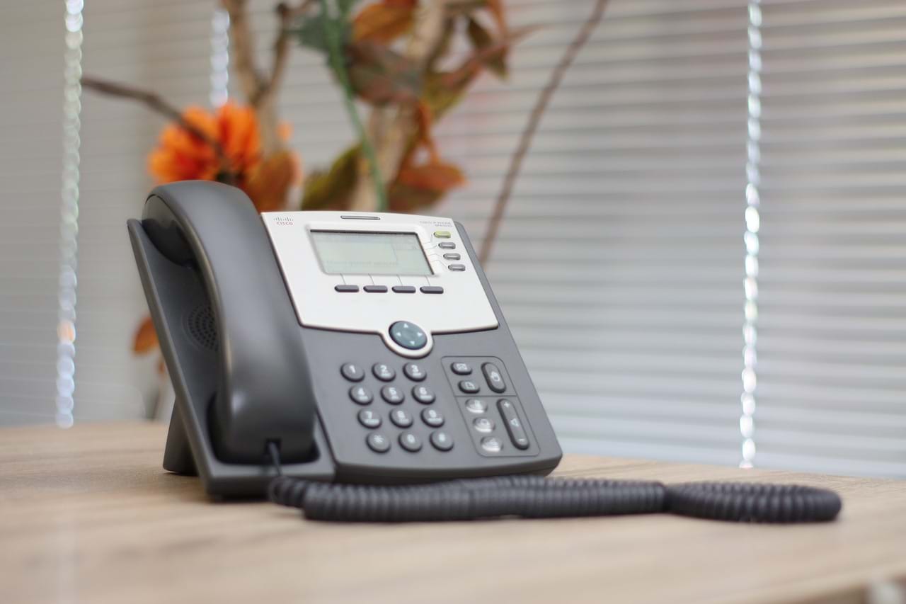 A black office phone with a digital display sits on a wooden desk. Behind it is a window with blinds and an out-of-focus plant with orange flowers.