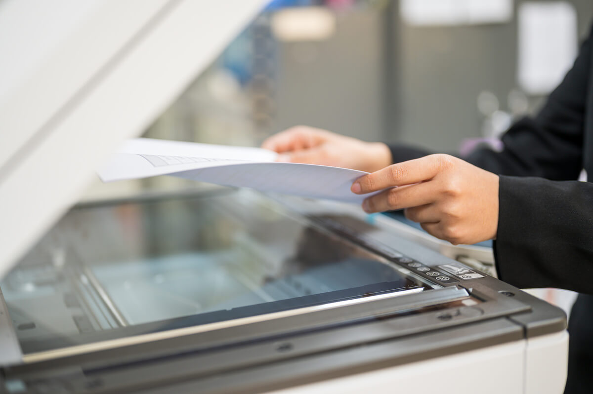 A person wearing a black suit places a document into a photocopier in an office setting, focusing on the task at hand.